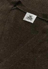 Sötétbarna kötött anyag, sima textúrával. Egy fehér címke fekete felirattal, a felső szélénél elhelyezve.