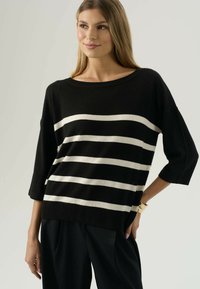 Donna che indossa un maglione nero con quattro strisce beige orizzontali, abbinato a pantaloni neri, in piedi con una mano su un fianco contro uno sfondo semplice.