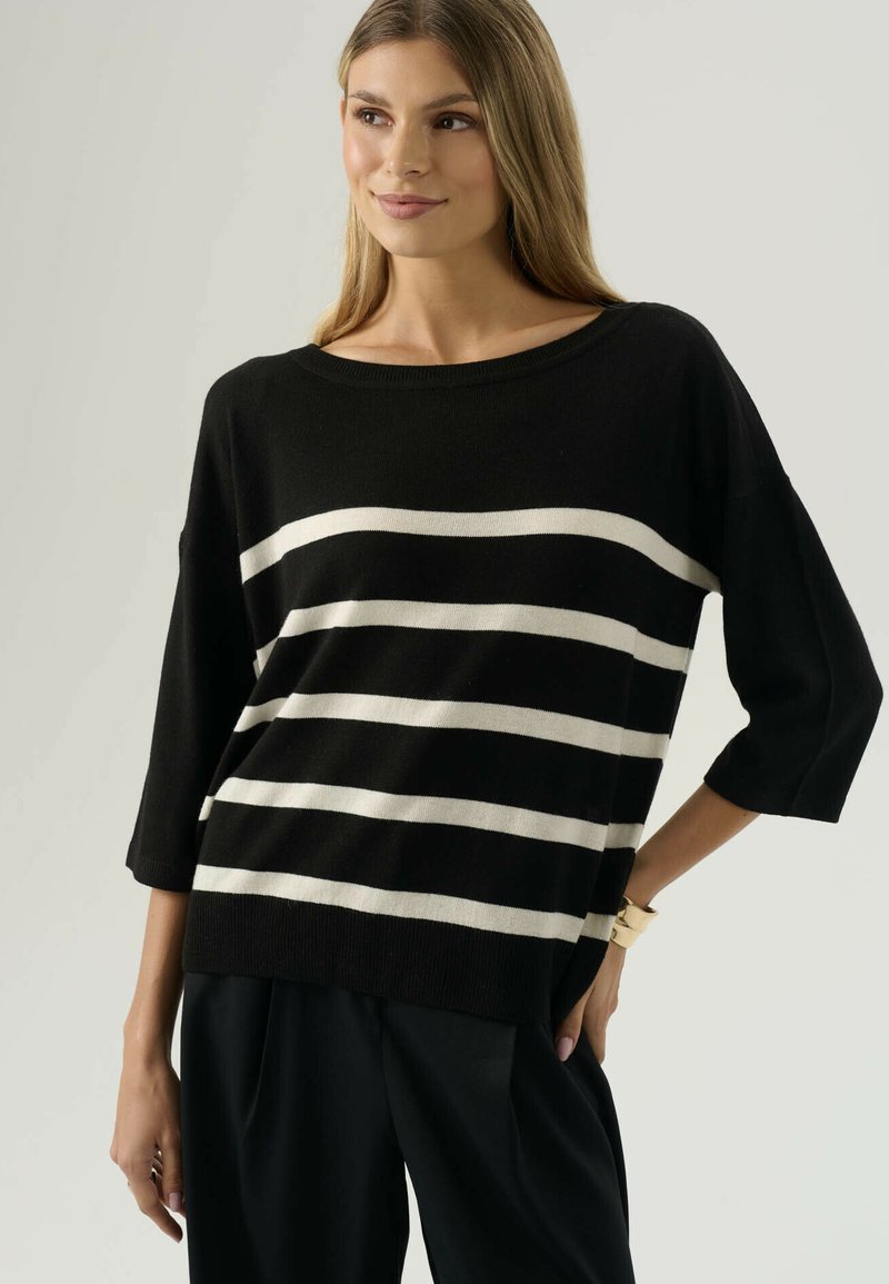 Donna che indossa un maglione nero con quattro strisce beige orizzontali, abbinato a pantaloni neri, in piedi con una mano su un fianco contro uno sfondo semplice.