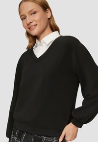 Czarny sweter z dekoltem w serek wykonany z miękkiej tkaniny, z prążkowanymi mankietami i dołem, o swobodnym kroju, noszony na białej koszuli z kołnierzykiem.