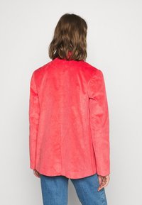 Červená kordurová sako se strukturovaným střihem, dlouhými rukávy a klopovým límcem. Viditelná je hladká textura a vertikální pruhování.