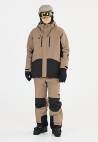 Beige och svart vattentät skidjacka med justerbar huva, fickor med dragkedja och matchande beige skidbyxor. Svarta skidstövlar fulländar outfiten.
