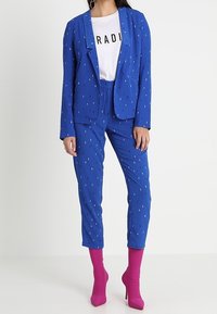 Modrý blejzr a kalhoty s bílými vertikálními pruhy, doplněné bílým tričkem s grafikou. Modelka má na sobě jasně růžové kotníkové boty.