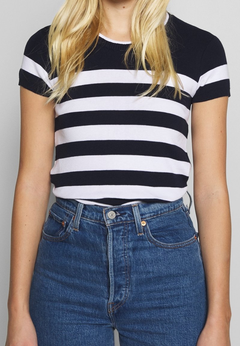 Námořnická a bílá pruhovaná blůza s krátkým rukávem a žebrovaným výstřihem, v kombinaci s modrými džíny s vysokým pasem. Tkanina se zdá být hladká a přiléhavá.