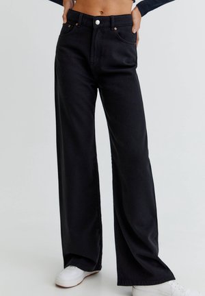 Jean noir taille haute à jambes larges porté avec des baskets blanches, montrant le ventre et les mains posées sur les hanches sur un fond uni.