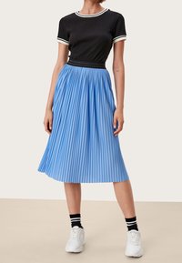 Donna che indossa un top nero a maniche corte, una gonna midi plissettata azzurra chiara, calze nere con strisce bianche e sneaker bianche, in piedi su un pavimento beige.