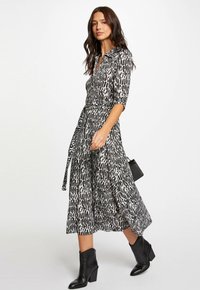 Zwart-witte gemusterde jurk met een kraag, driekwart mouwen, een ceintuur in de taille en een vloeiende rok. G gedragen met zwarte enkellaarzen.