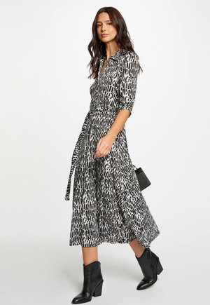 Schwarz-weiß gemustertes Kleid mit Kragen, Dreiviertelärmeln, tailliertem Bund und fließendem Rock. Getragen mit schwarzen Knöchelstiefeln.