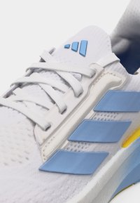 Λευκό αθλητικό παπούτσι με ανοιχτό μπλε ρίγες Adidas, λευκά κορδόνια και κίτρινη λεπτομέρεια στη λευκή σόλα.
