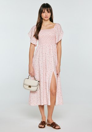 Kobieta w różowej sukience midi w kwiaty z bufkami, z rozcięciem na udzie, trzyma kremową torebkę i nosi brązowe sandały wsuwane na szarym tle.