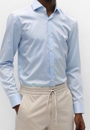 Man wearing light blue buttoned shirt tucked into beige drawstring pants with hands in pockets, shown from neck to hips.