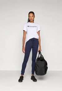 Moteris stovi, vilkėdama baltą Champion marškinėlį, tamsiai mėlynas sporto kelnes, juodus sportbačius, laikydama didelę juodą sporto maišelį prieš paprastą fono.