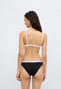 Μαύρα βαμβακερά εσώρουχα με λευκή μέση που φέρει το λογότυπο "TOMMY HILFIGER"· συνοδεύονται από λευκό σουτιέν, με τους ιμάντες ορατούς στους ώμους.