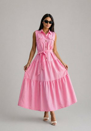 Femme portant une robe maxi rose sans manches boutonnée avec ceinture, lunettes de soleil noires et talons blancs sur fond gris uni.