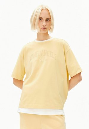Jeune personne aux cheveux courts et blonds portant un t-shirt ample jaune clair avec un texte léger "ARMEDANGELS" et un pantalon assorti.
