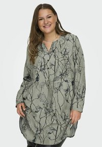Tunique à manches longues verte avec des imprimés noirs à base de plantes, dotée d'un col et d'une patte de boutonnage dissimulée. Tissu lisse et fluide.