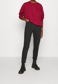 Sudadera roja, jeans ajustados negros y botas negras con cordones. La sudadera tiene un cuello redondo y un corte relajado. Los jeans tienen puños enrollados y un diseño minimalista.