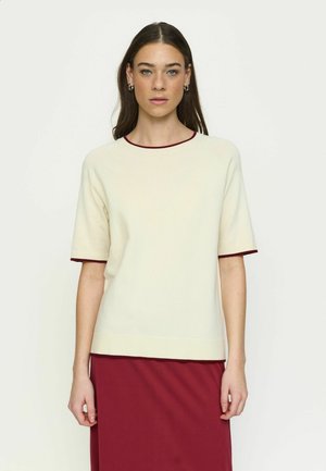 Naine, kellel on pikad tumejuuksed, kannab kreemja lühikeste varrukatega kampsunit marooni servadega ja sobivat marooni seelikut, seisab ühevärvilise tausta ees.