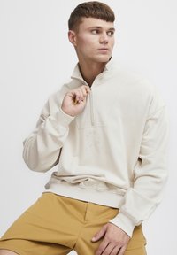 Sudadera con cremallera de color beige claro y cuello alto, con textura acanalada, que presenta un logo circular en el pecho y puños elásticos.