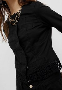 Mujer que lleva una chaqueta negra entallada con encaje en el dobladillo y una collar de cadenas de oro en capas.