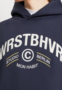 Sudadera azul marino con texto bordado en blanco y amarillo: "WRSTBHVR STUDIO BERLIN MON HABIT" y un logo circular. Textura de tela suave.