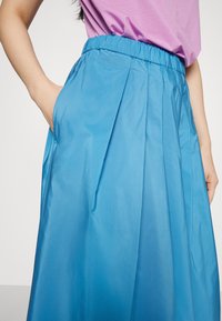 Jupe plissée bleue avec une ceinture élastique et des poches latérales ; texture lisse, tissu léger et finition légèrement brillante.