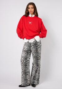 Punainen collegepaita, jossa on kirjailtu rusettimerkki, yhdistettynä leveälahkeisiin leopardi-kuvioisiin paljettihousuihin mustavalkoisina. Mustat jalkineet näkyvissä.