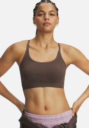Fit woman with braided hair wears brown sports bra and light purple pants, standing with hands on hips against a plain background.