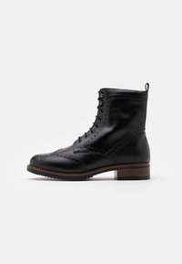 Botín negro de cuero con cordones, con perforaciones de brogue, tacón apilado marrón y una lengüeta en la parte trasera sobre un fondo blanco.