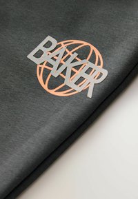 Dunkelgrauer Textilstoff mit glatter Struktur, der ein deutlich sichtbares hellgraues "BAKER"-Logo und ein orangefarbenes rundes Design hinter dem Schriftzug zeigt.