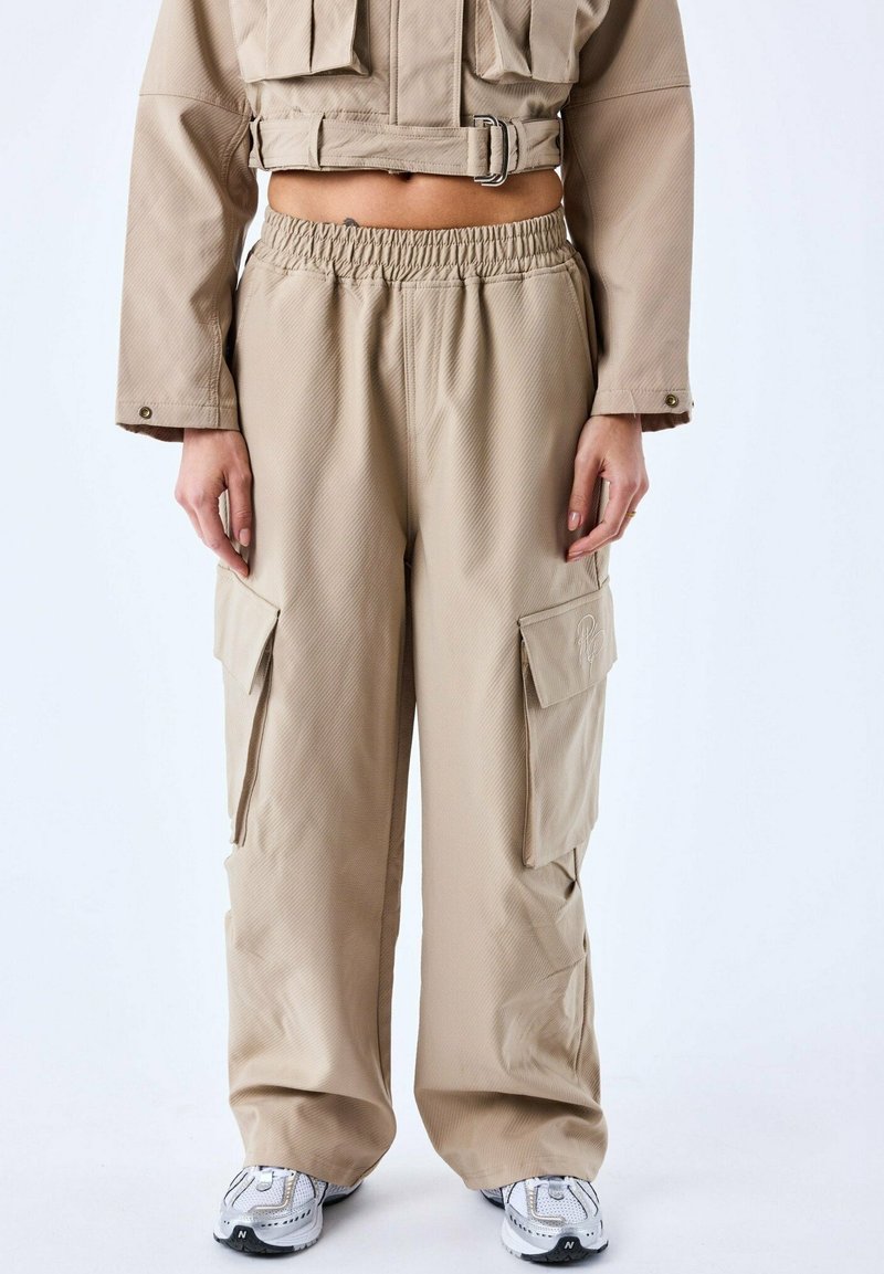 Pantalons cargo beiges avec une taille élastique, grandes poches latérales et tissu texturé. Associés à des baskets blanches avec des accents gris.