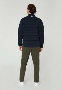 Marinblå pufferjacka med horisontell sömnad, hög krage och logodetalj på ryggen. Parat med olivgröna byxor och vita sneakers.