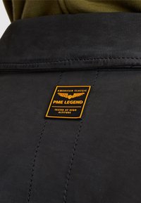 Zwarte leren jas met een opvallende gele en zwarte merkpatch met vleugels, gedetailleerde stiksels langs de schoudernaad.