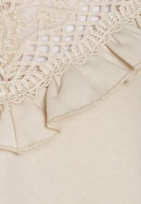 Beżowa tkanina z teksturowanym, kwiatowym haftem i falbaną na krawędzi; gładka faktura o lekkim, przewiewnym wyglądzie.