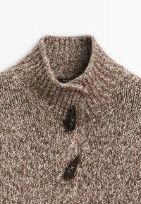 Hnědý pletený svetr s vysokým límcem, s texturovaným vzorem a dvěma tmavými knoflíky na zapínání u krku.
