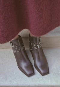 Hnědé kožené kotníkové boty s špičatou špičkou, zdobené křížovými pásky s kovovými oky. Hladký povrch, minimalistický design.