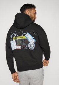 Μαύρη κουκούλα με γραφικό αναχώρησης αεροδρομίου στην πλάτη, συμπεριλαμβανομένων των εισιτηρίων επιβίβασης και φυσαλίδων σε μπλε και λευκό.