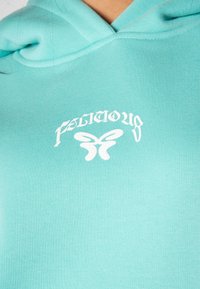Félkapucnis pulóver világos teal színben, fehér grafikai szöveggel és lepkemotívummal a mellrészen. Pamut keverék anyag, sima tapintás.