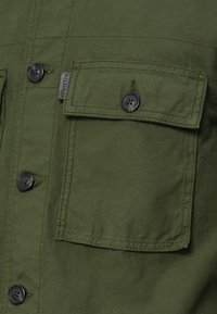 Oliivroheline kangas, millel on nööpidega klapiga tasku ja mustad nööbid riidel, ning väikene kangasilti.