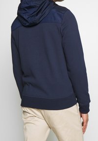 Sudadera azul marino con capucha, que presenta una textura suave y un tejido brillante en los hombros. Puños y dobladillo elásticos.
