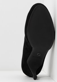 Chaussure à talon haut en daim noir avec un bout pointu et un talon fin. La semelle est lisse et plate, avec un logo estampillé près du talon.