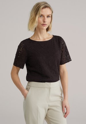 Žena s krátkymi blond vlasmi, oblečená v čiernom čipkovanom tričku s krátkym rukávom a svetlo béžových vysokých nohaviciach, stojaca s jednou rukou v vrecku.