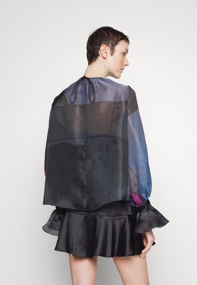 Blouse fluide et transparente en bleu foncé et noir, avec des manches longues et des poignets à volants. Le dos présente une fermeture à cordon et un design en tissus superposés.