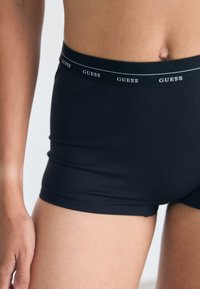 Boxer-briefs noirs en tissu extensible avec une large ceinture élastique arborant le logo "GUESS" répété en blanc. Texture lisse et design ajusté.