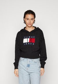 Crna kabelna kapa s pletivom, skraćena kapuljača s prednjom oznakom u crvenoj, bijeloj i mornarskoj plavoj boji. Uparena s svijetloplavim hlačama visokog struka.