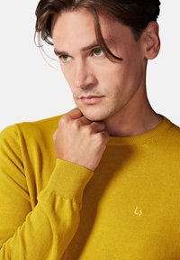 Żółty sweter z dzianiny z okrągłym dekoltem, ściągaczami na rękawach i subtelnym detalem z logo na piersi. Gładka struktura, dopasowany krój.