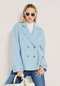 Frau in einem übergroßen hellblauen doppelt geknöpften Mantel, weißem Rollkragenpullover, weißen Sonnenbrillen und blauen Jeans, die eine gesteppte cremefarbene Handtasche hält und große Ringe trägt.