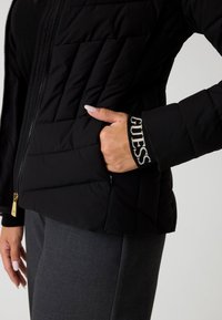Sort quiltet jakke med lynlås, sidelommer og ribbede manchetter med "GUESS" branding i cremefarve. Glat tekstur.