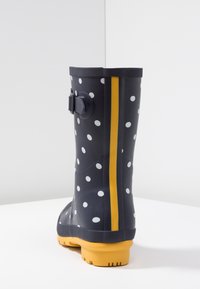 Musta pilkullinen kuminen sadekenkä, jossa on keltainen raita ja sivuolkain yksityiskohta. Raekuvioitu pohja ja sileä päällismateriaali.