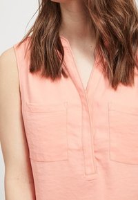 Blouse corail sans manches avec un décolleté en V, deux poches poitrine et des détails de couture subtils. Tissu doux avec une texture lisse.